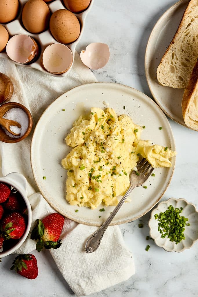 Completed scrambled egg recipe using cottage cheese on a plate with a fork, with eggshells, a plate of toast, a small dish of chives and salt, and a small dish of strawberries nearby