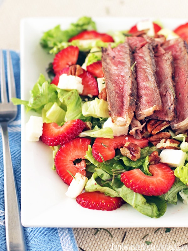 Sliced steak on a plate of salad greens with sliced strawberries.