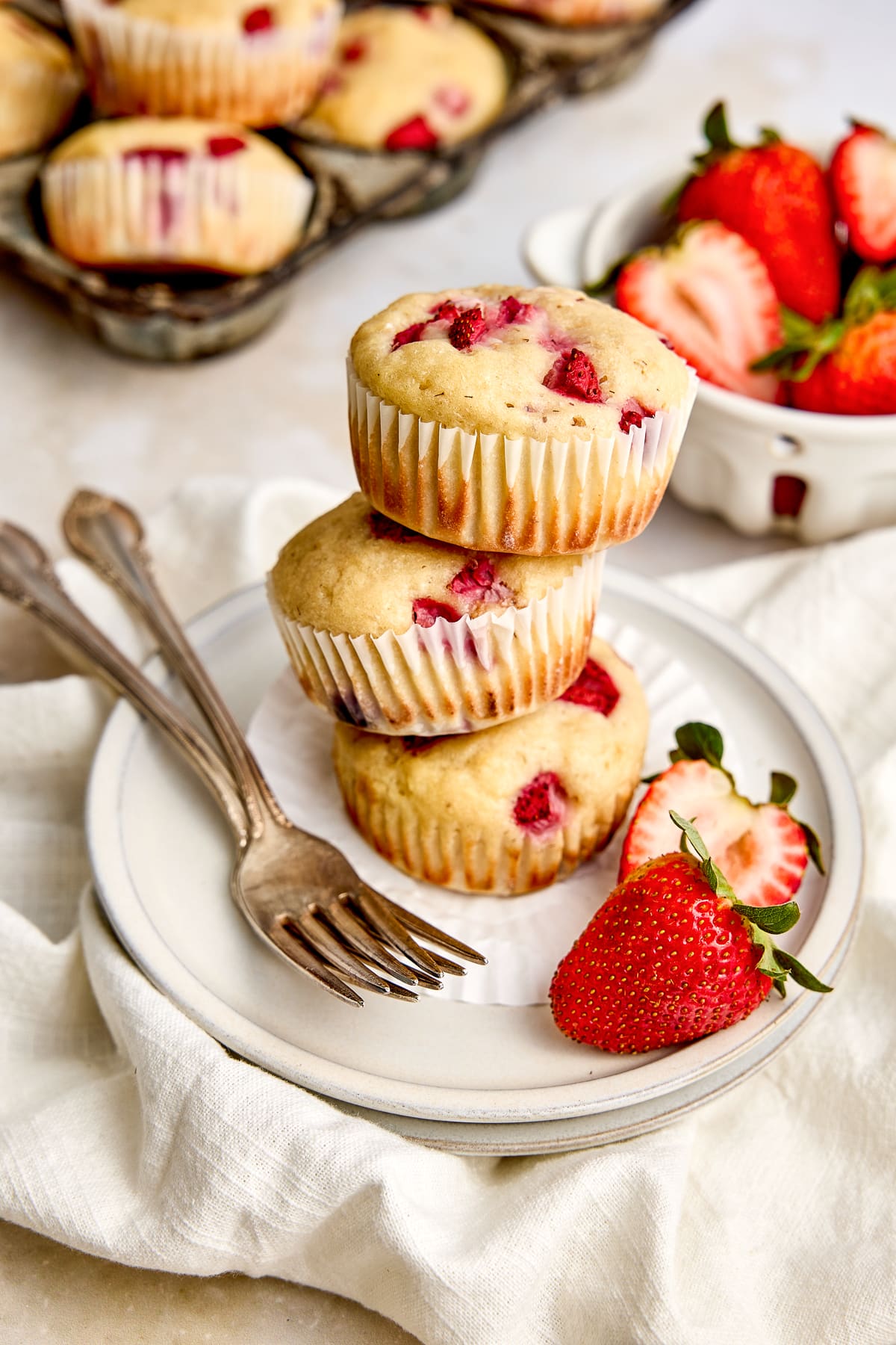 Three protein muffins with fruit stacked on a plate with two forks and a strawberry, with a bowl of fresh strawberries in the background