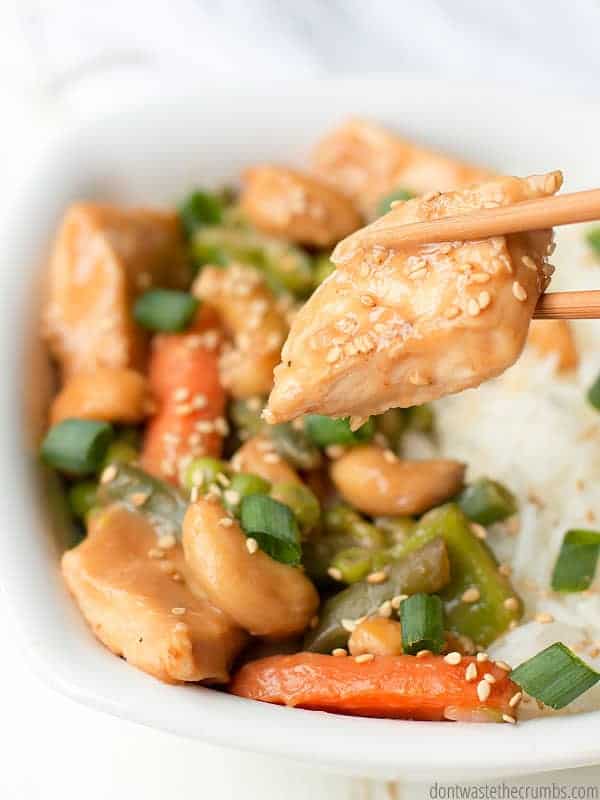 Chopsticks holding a piece of cashew chicken, with a bowl of cashew chicken, veggies, and white rice in the background